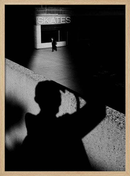 A high-contrast, black and white street photograph where the large shadow of the photographer is cast on a concrete barrier in the foreground. In the background, a lone person walks through a spotlit area of sidewalk in front of a building with an illuminated sign that says 'SKATES'. Poster