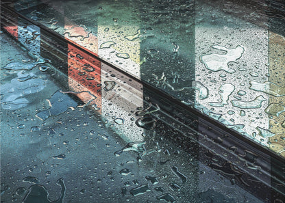 An abstract close-up photograph of a wet, dark surface, possibly a car, covered in raindrops, with a prominent black diagonal line. Colorful vertical stripes of red, cream, and green are reflected on the glossy, wet surface. The artwork is presented in a silver frame. Decor