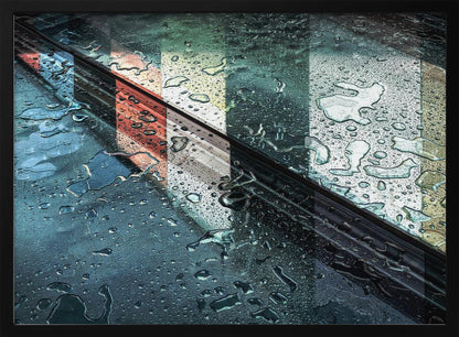 An abstract close-up photograph of a wet, dark surface, possibly a car, covered in raindrops, with a prominent black diagonal line. Colorful vertical stripes of red, cream, and green are reflected on the glossy, wet surface. The artwork is presented in a silver frame. Decor