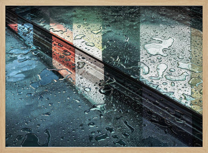 An abstract close-up photograph of a wet, dark surface, possibly a car, covered in raindrops, with a prominent black diagonal line. Colorful vertical stripes of red, cream, and green are reflected on the glossy, wet surface. The artwork is presented in a silver frame. Decor