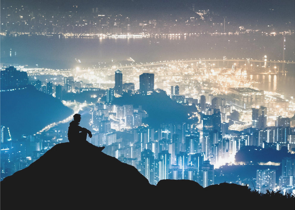 A silhouette of a person sitting on a hilltop, gazing out over a vast, illuminated city skyline at night. The city lights glow in shades of blue and gold against the dark landscape, with a body of water in the distance. Decor
