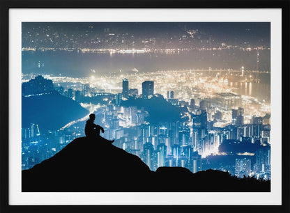 A silhouette of a person sitting on a hilltop, gazing out over a vast, illuminated city skyline at night. The city lights glow in shades of blue and gold against the dark landscape, with a body of water in the distance. Decor