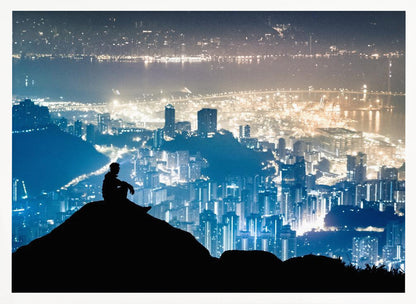 A silhouette of a person sitting on a hilltop, gazing out over a vast, illuminated city skyline at night. The city lights glow in shades of blue and gold against the dark landscape, with a body of water in the distance. Decor