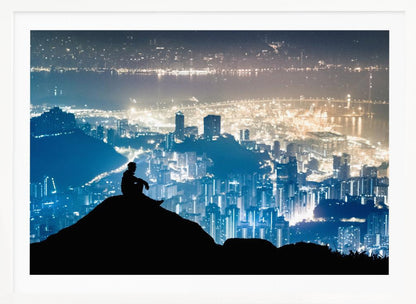 A silhouette of a person sitting on a hilltop, gazing out over a vast, illuminated city skyline at night. The city lights glow in shades of blue and gold against the dark landscape, with a body of water in the distance. Decor