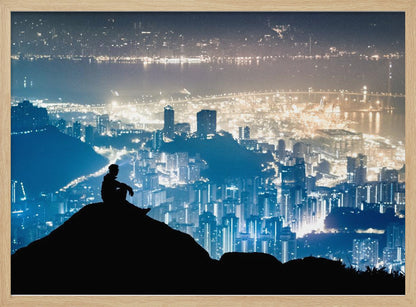 A silhouette of a person sitting on a hilltop, gazing out over a vast, illuminated city skyline at night. The city lights glow in shades of blue and gold against the dark landscape, with a body of water in the distance. Decor