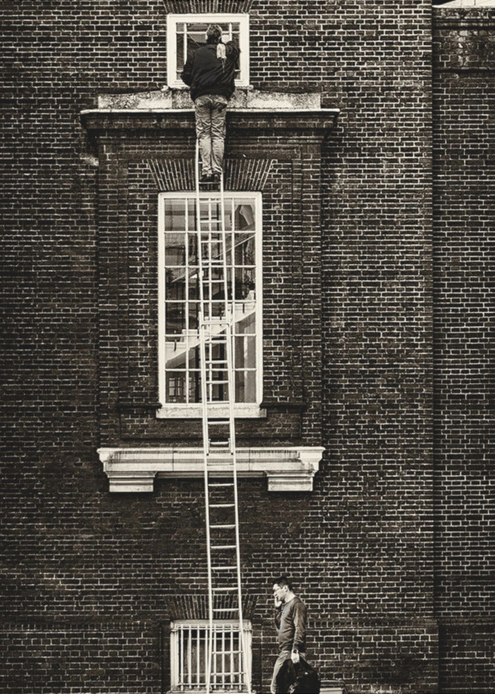 A striking black and white photograph of a man cleaning a small window high up on a brick building, standing precariously at the top of a very long ladder. Below, another man walks past on the street, seemingly unaware of the scene above. Artwork