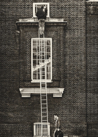 A striking black and white photograph of a man cleaning a small window high up on a brick building, standing precariously at the top of a very long ladder. Below, another man walks past on the street, seemingly unaware of the scene above. Artwork