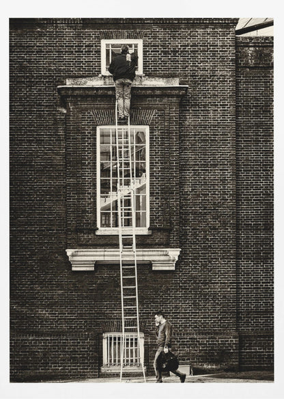A striking black and white photograph of a man cleaning a small window high up on a brick building, standing precariously at the top of a very long ladder. Below, another man walks past on the street, seemingly unaware of the scene above. Artwork