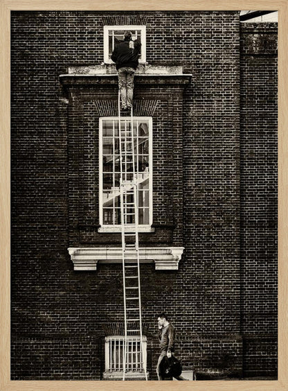 A striking black and white photograph of a man cleaning a small window high up on a brick building, standing precariously at the top of a very long ladder. Below, another man walks past on the street, seemingly unaware of the scene above. Artwork