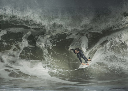 A dramatic overhead photograph of a surfer in a black wetsuit riding a white surfboard on a large, dark, foamy wave. The scene is captured in a moody, almost monochromatic tone and is presented within a silver picture frame. Artwork
