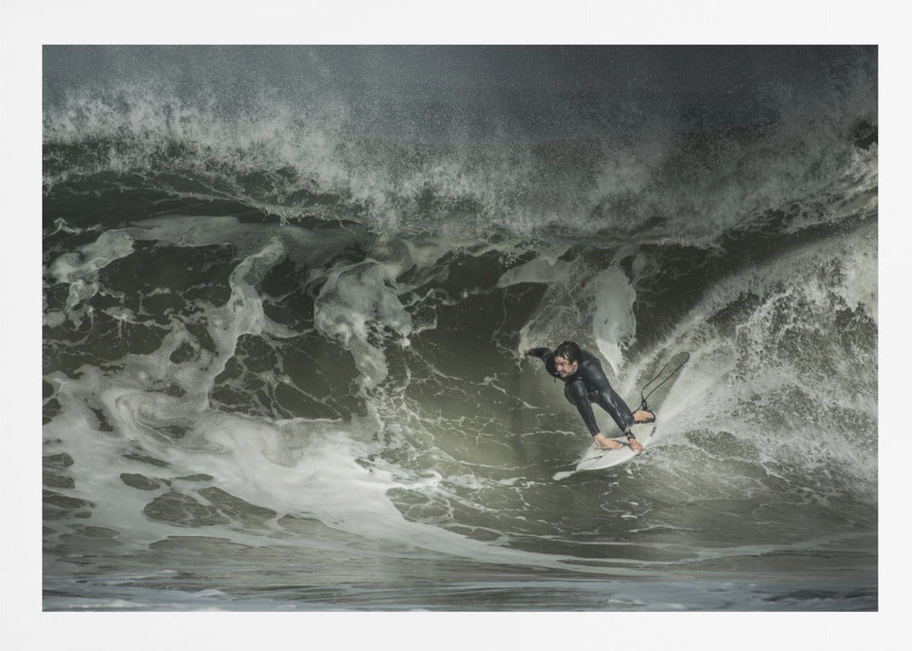 A dramatic overhead photograph of a surfer in a black wetsuit riding a white surfboard on a large, dark, foamy wave. The scene is captured in a moody, almost monochromatic tone and is presented within a silver picture frame. Artwork