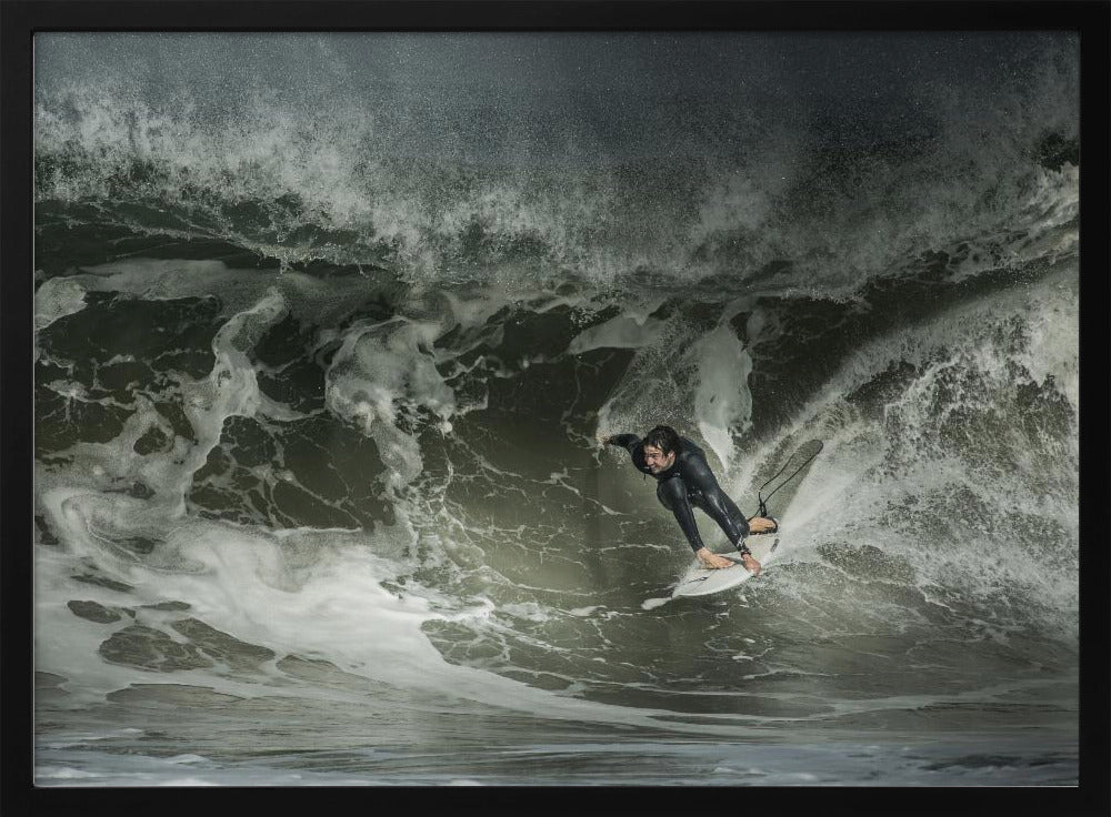 A dramatic overhead photograph of a surfer in a black wetsuit riding a white surfboard on a large, dark, foamy wave. The scene is captured in a moody, almost monochromatic tone and is presented within a silver picture frame. Artwork