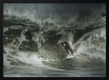 A dramatic overhead photograph of a surfer in a black wetsuit riding a white surfboard on a large, dark, foamy wave. The scene is captured in a moody, almost monochromatic tone and is presented within a silver picture frame. Artwork