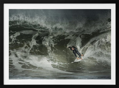 A dramatic overhead photograph of a surfer in a black wetsuit riding a white surfboard on a large, dark, foamy wave. The scene is captured in a moody, almost monochromatic tone and is presented within a silver picture frame. Artwork