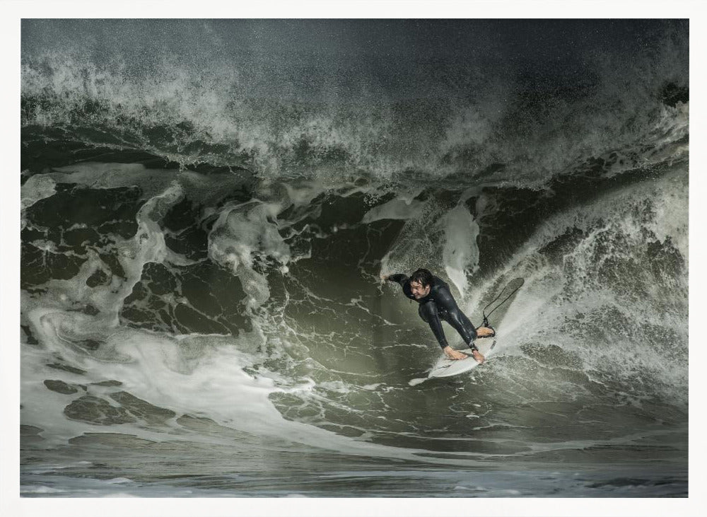 A dramatic overhead photograph of a surfer in a black wetsuit riding a white surfboard on a large, dark, foamy wave. The scene is captured in a moody, almost monochromatic tone and is presented within a silver picture frame. Artwork