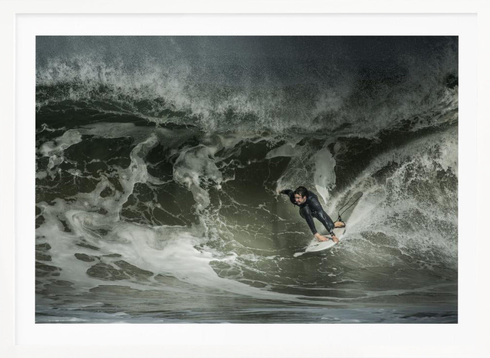 A dramatic overhead photograph of a surfer in a black wetsuit riding a white surfboard on a large, dark, foamy wave. The scene is captured in a moody, almost monochromatic tone and is presented within a silver picture frame. Artwork