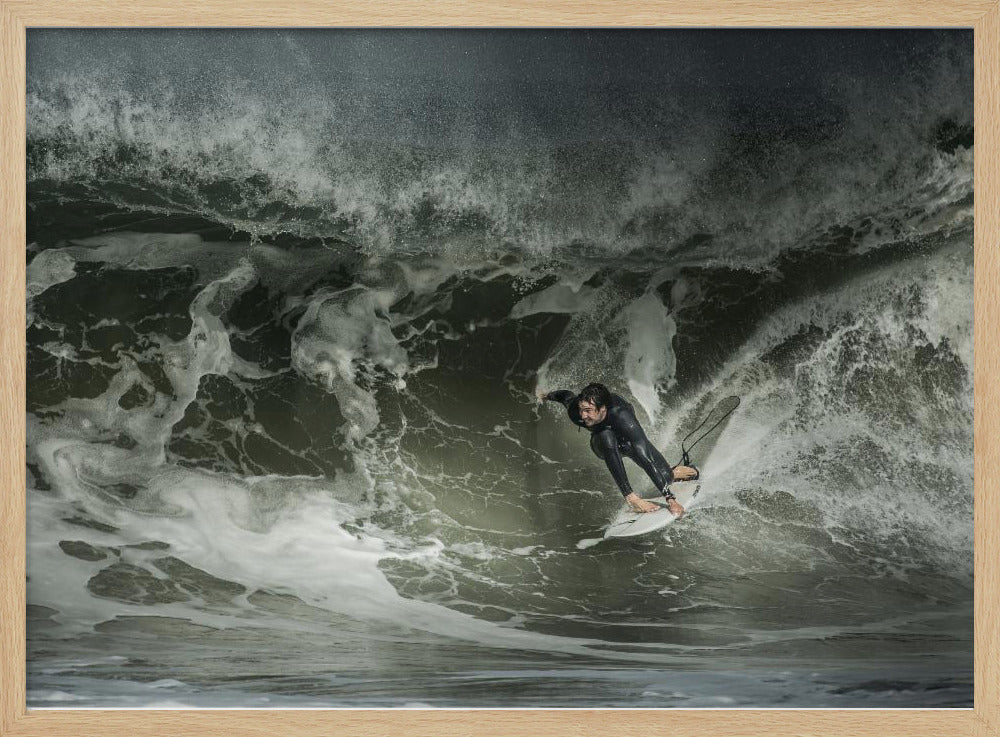 A dramatic overhead photograph of a surfer in a black wetsuit riding a white surfboard on a large, dark, foamy wave. The scene is captured in a moody, almost monochromatic tone and is presented within a silver picture frame. Artwork