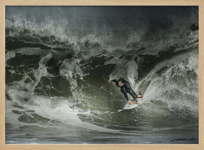A dramatic overhead photograph of a surfer in a black wetsuit riding a white surfboard on a large, dark, foamy wave. The scene is captured in a moody, almost monochromatic tone and is presented within a silver picture frame. Artwork