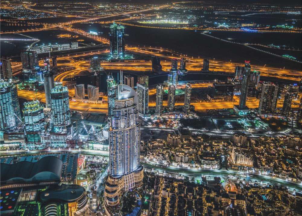 An aerial nighttime photograph of a sprawling modern city, possibly Dubai, with brightly lit skyscrapers and a complex network of illuminated highways creating a vibrant contrast against the dark landscape, all enclosed in a silver frame. Decor