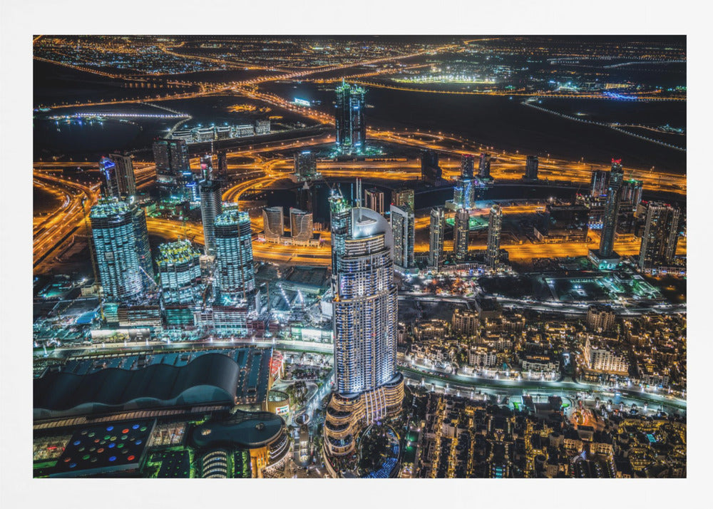 An aerial nighttime photograph of a sprawling modern city, possibly Dubai, with brightly lit skyscrapers and a complex network of illuminated highways creating a vibrant contrast against the dark landscape, all enclosed in a silver frame. Decor