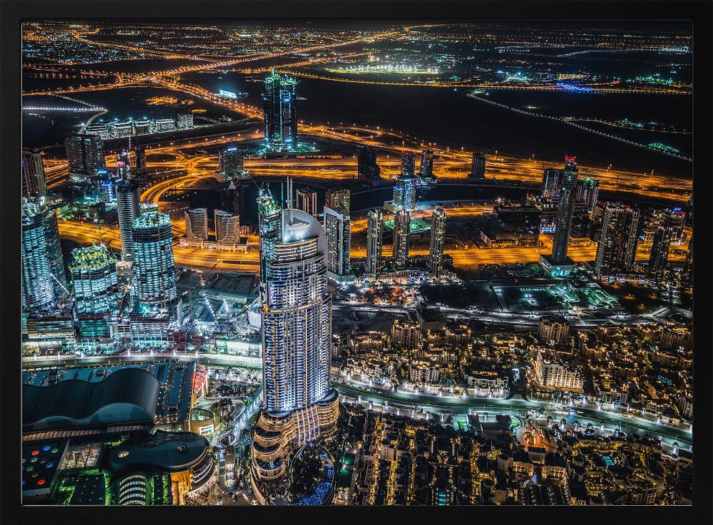 An aerial nighttime photograph of a sprawling modern city, possibly Dubai, with brightly lit skyscrapers and a complex network of illuminated highways creating a vibrant contrast against the dark landscape, all enclosed in a silver frame. Decor