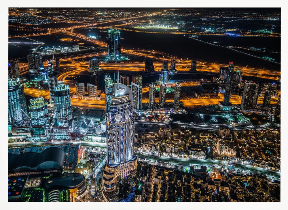 An aerial nighttime photograph of a sprawling modern city, possibly Dubai, with brightly lit skyscrapers and a complex network of illuminated highways creating a vibrant contrast against the dark landscape, all enclosed in a silver frame. Decor