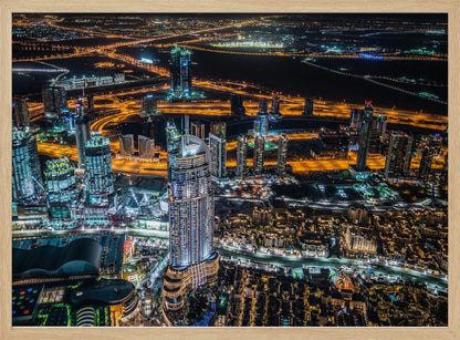 An aerial nighttime photograph of a sprawling modern city, possibly Dubai, with brightly lit skyscrapers and a complex network of illuminated highways creating a vibrant contrast against the dark landscape, all enclosed in a silver frame. Decor