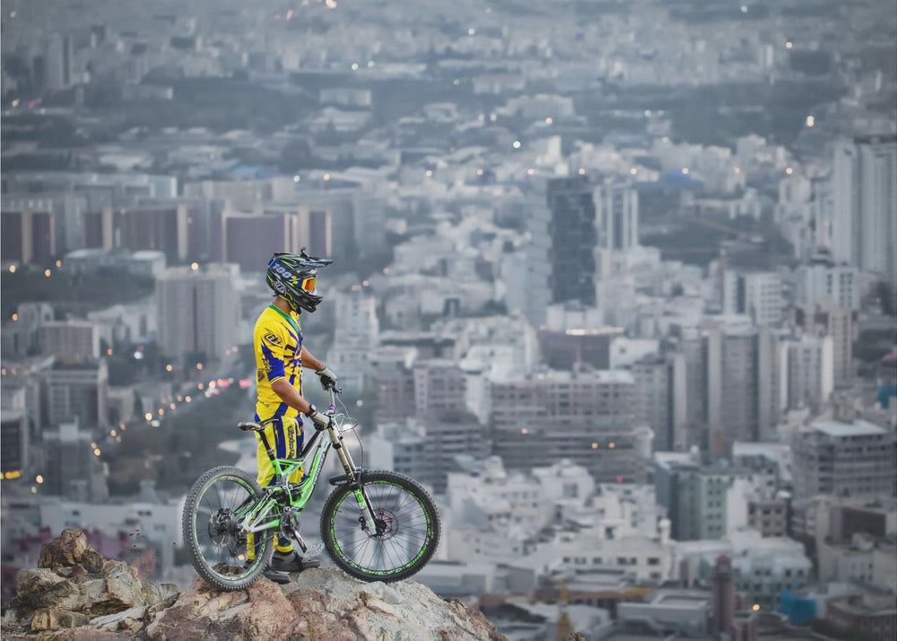 A mountain biker in a yellow and blue uniform stands with a green and white full-suspension bike on a rocky precipice, looking out over a vast, hazy cityscape that stretches into the distance. Print