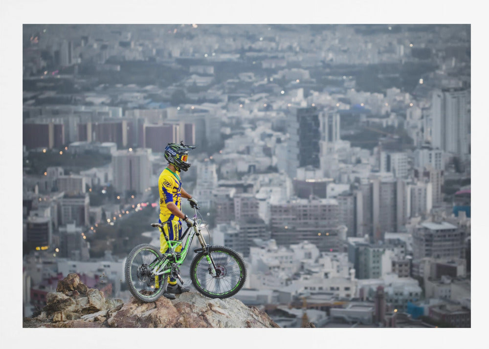 A mountain biker in a yellow and blue uniform stands with a green and white full-suspension bike on a rocky precipice, looking out over a vast, hazy cityscape that stretches into the distance. Print