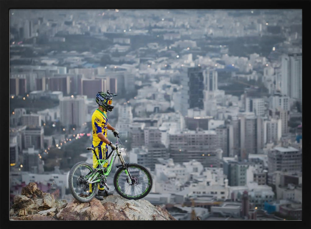 A mountain biker in a yellow and blue uniform stands with a green and white full-suspension bike on a rocky precipice, looking out over a vast, hazy cityscape that stretches into the distance. Print