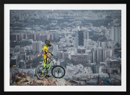 A mountain biker in a yellow and blue uniform stands with a green and white full-suspension bike on a rocky precipice, looking out over a vast, hazy cityscape that stretches into the distance. Print