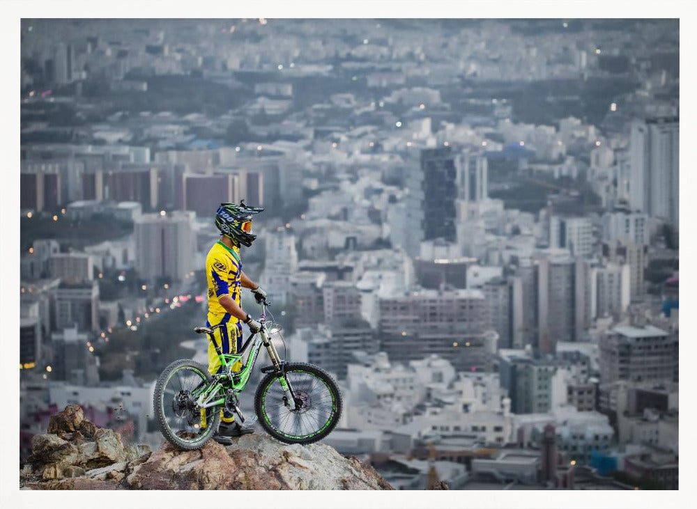 A mountain biker in a yellow and blue uniform stands with a green and white full-suspension bike on a rocky precipice, looking out over a vast, hazy cityscape that stretches into the distance. Print