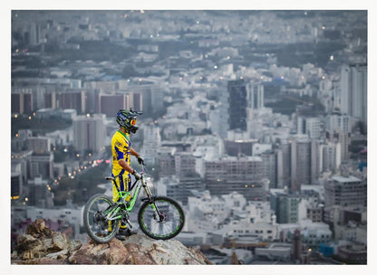 A mountain biker in a yellow and blue uniform stands with a green and white full-suspension bike on a rocky precipice, looking out over a vast, hazy cityscape that stretches into the distance. Print