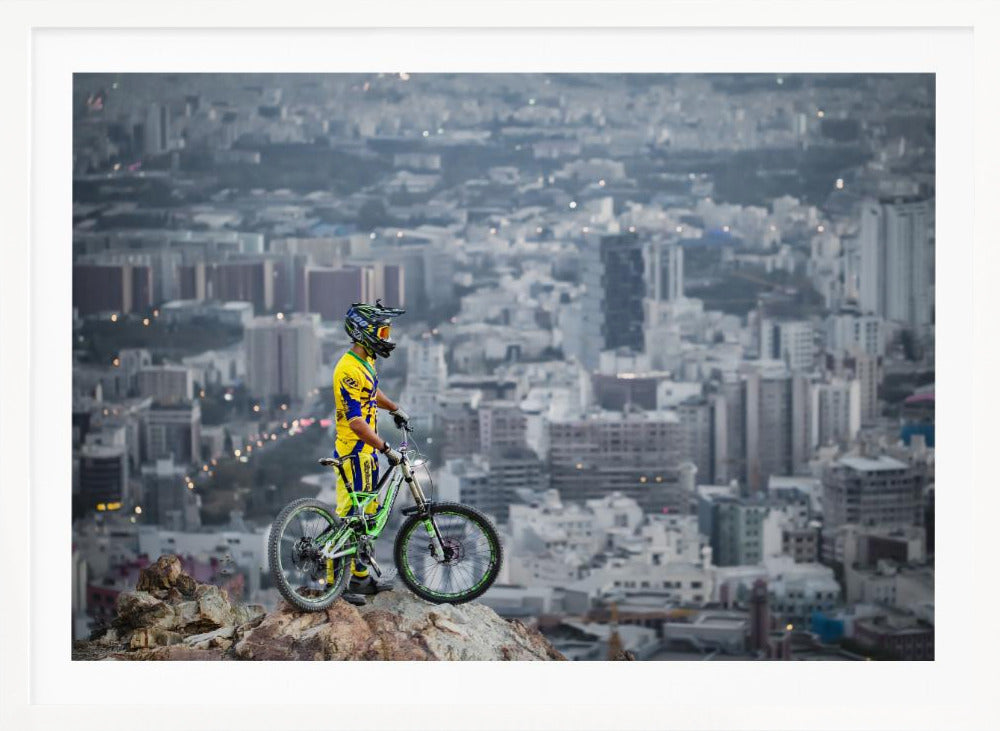 A mountain biker in a yellow and blue uniform stands with a green and white full-suspension bike on a rocky precipice, looking out over a vast, hazy cityscape that stretches into the distance. Print