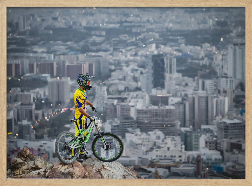 A mountain biker in a yellow and blue uniform stands with a green and white full-suspension bike on a rocky precipice, looking out over a vast, hazy cityscape that stretches into the distance. Print