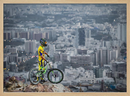 A mountain biker in a yellow and blue uniform stands with a green and white full-suspension bike on a rocky precipice, looking out over a vast, hazy cityscape that stretches into the distance. Print
