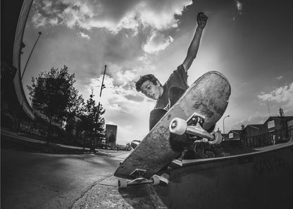 A dynamic, low-angle, black and white photograph of a young man skateboarding. He is balancing on the edge of a ramp with his skateboard angled up towards the viewer. He has one arm raised in the air with a clenched fist. The shot is taken with a fisheye lens, curving the horizon against a dramatic cloudy sky in an urban setting. The image is presented within a silver frame. Decor