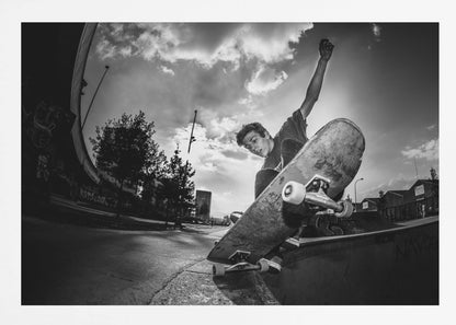 A dynamic, low-angle, black and white photograph of a young man skateboarding. He is balancing on the edge of a ramp with his skateboard angled up towards the viewer. He has one arm raised in the air with a clenched fist. The shot is taken with a fisheye lens, curving the horizon against a dramatic cloudy sky in an urban setting. The image is presented within a silver frame. Decor