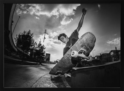 A dynamic, low-angle, black and white photograph of a young man skateboarding. He is balancing on the edge of a ramp with his skateboard angled up towards the viewer. He has one arm raised in the air with a clenched fist. The shot is taken with a fisheye lens, curving the horizon against a dramatic cloudy sky in an urban setting. The image is presented within a silver frame. Decor