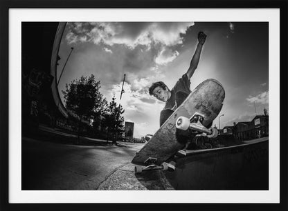 A dynamic, low-angle, black and white photograph of a young man skateboarding. He is balancing on the edge of a ramp with his skateboard angled up towards the viewer. He has one arm raised in the air with a clenched fist. The shot is taken with a fisheye lens, curving the horizon against a dramatic cloudy sky in an urban setting. The image is presented within a silver frame. Decor