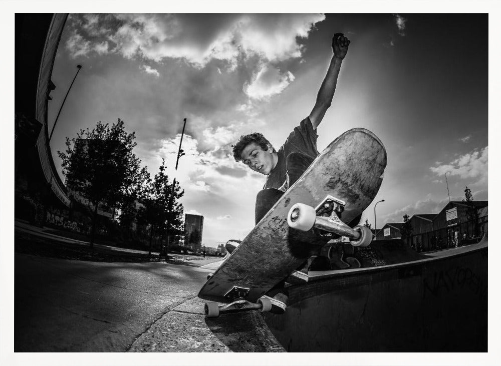 A dynamic, low-angle, black and white photograph of a young man skateboarding. He is balancing on the edge of a ramp with his skateboard angled up towards the viewer. He has one arm raised in the air with a clenched fist. The shot is taken with a fisheye lens, curving the horizon against a dramatic cloudy sky in an urban setting. The image is presented within a silver frame. Decor