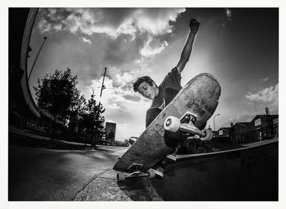 A dynamic, low-angle, black and white photograph of a young man skateboarding. He is balancing on the edge of a ramp with his skateboard angled up towards the viewer. He has one arm raised in the air with a clenched fist. The shot is taken with a fisheye lens, curving the horizon against a dramatic cloudy sky in an urban setting. The image is presented within a silver frame. Decor