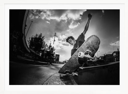 A dynamic, low-angle, black and white photograph of a young man skateboarding. He is balancing on the edge of a ramp with his skateboard angled up towards the viewer. He has one arm raised in the air with a clenched fist. The shot is taken with a fisheye lens, curving the horizon against a dramatic cloudy sky in an urban setting. The image is presented within a silver frame. Decor