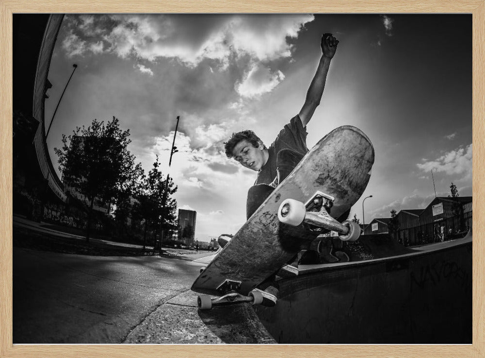 A dynamic, low-angle, black and white photograph of a young man skateboarding. He is balancing on the edge of a ramp with his skateboard angled up towards the viewer. He has one arm raised in the air with a clenched fist. The shot is taken with a fisheye lens, curving the horizon against a dramatic cloudy sky in an urban setting. The image is presented within a silver frame. Decor