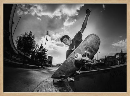 A dynamic, low-angle, black and white photograph of a young man skateboarding. He is balancing on the edge of a ramp with his skateboard angled up towards the viewer. He has one arm raised in the air with a clenched fist. The shot is taken with a fisheye lens, curving the horizon against a dramatic cloudy sky in an urban setting. The image is presented within a silver frame. Decor