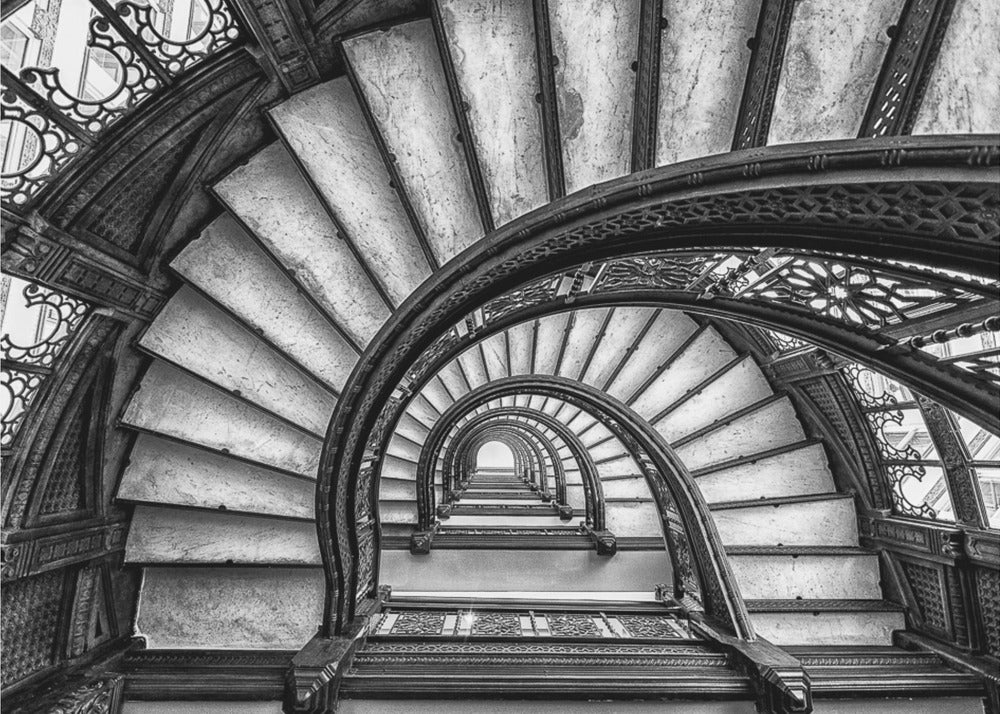 A dramatic black and white photograph looking up from the bottom of an ornate spiral staircase. The dark, intricate ironwork of the railings contrasts with the light stone steps, creating a mesmerizing, repeating pattern that spirals towards a bright light at the top. The image is presented within a silver-colored frame. Decor