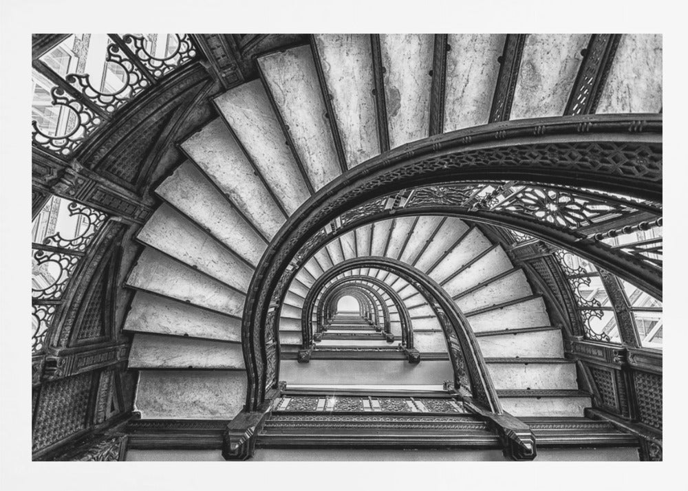 A dramatic black and white photograph looking up from the bottom of an ornate spiral staircase. The dark, intricate ironwork of the railings contrasts with the light stone steps, creating a mesmerizing, repeating pattern that spirals towards a bright light at the top. The image is presented within a silver-colored frame. Decor
