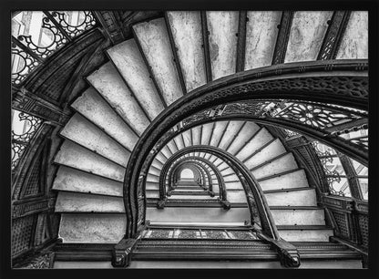 A dramatic black and white photograph looking up from the bottom of an ornate spiral staircase. The dark, intricate ironwork of the railings contrasts with the light stone steps, creating a mesmerizing, repeating pattern that spirals towards a bright light at the top. The image is presented within a silver-colored frame. Decor