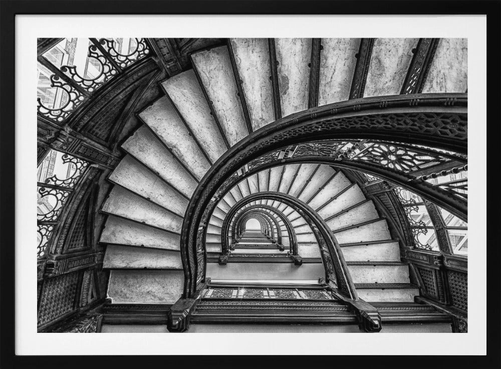 A dramatic black and white photograph looking up from the bottom of an ornate spiral staircase. The dark, intricate ironwork of the railings contrasts with the light stone steps, creating a mesmerizing, repeating pattern that spirals towards a bright light at the top. The image is presented within a silver-colored frame. Decor