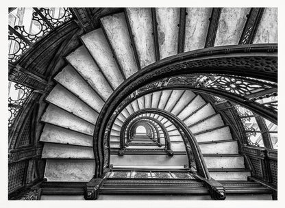 A dramatic black and white photograph looking up from the bottom of an ornate spiral staircase. The dark, intricate ironwork of the railings contrasts with the light stone steps, creating a mesmerizing, repeating pattern that spirals towards a bright light at the top. The image is presented within a silver-colored frame. Decor